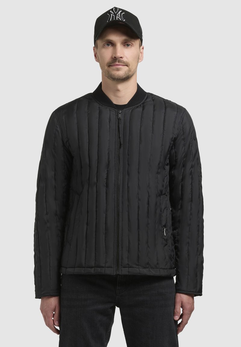 Black quilted jacket with vertical stitching and a front zipper. Ribbed collar and side pockets. Model paired with a black cap.