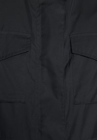 Svart tygjacka med två stora framfickor med klaffstängning och en slät, matt yta. Minimalistisk design utan synliga mönster.