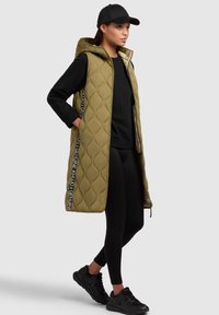 Gilet lungo quiltato verde oliva con cappuccio, abbinato a una felpa nera e leggings. Sneaker nere completano l'outfit.