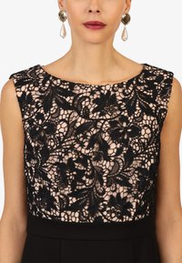 Un haut en dentelle noire avec un motif floral sur une doublure nude, associé à une jupe noire unie. Présente un col bateau et un design sans manches.