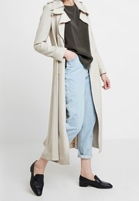 Trench-coat beige à double boutonnage, porté sur une chemise foncée et un jean bleu clair avec revers, associé à des mocassins noirs.