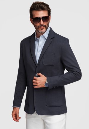 Mężczyzna w ciemnogranatowej marynarce, jasnobłękitnej koszuli w paski, białych spodniach, dużych okularach przeciwsłonecznych i zegarku na rękę, poprawia guzik marynarki.