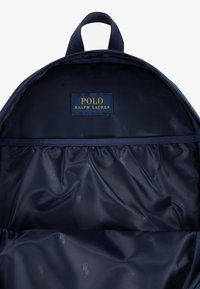 Vnitřek batohu tmavě modré barvy s hladou, texturovanou podšívkou. Obsahuje zlatou nášivku "POLO RALPH LAUREN" na tmavém látkovém pozadí.