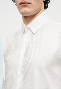 Άνδρας που φοράει λευκό πουκάμισο με κουμπιά, με διακριτικό υφασμάτινο σχέδιο και κλασικό μυτερό γιακά, εμφανιζόμενος από τον λαιμό έως το στήθος.