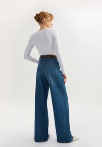 Jeans denim lebar di biru sedang, dilengkapi dengan dua saku belakang dan sabuk cokelat tua. Atasan berupa kemeja putih lengan panjang yang pas badan.