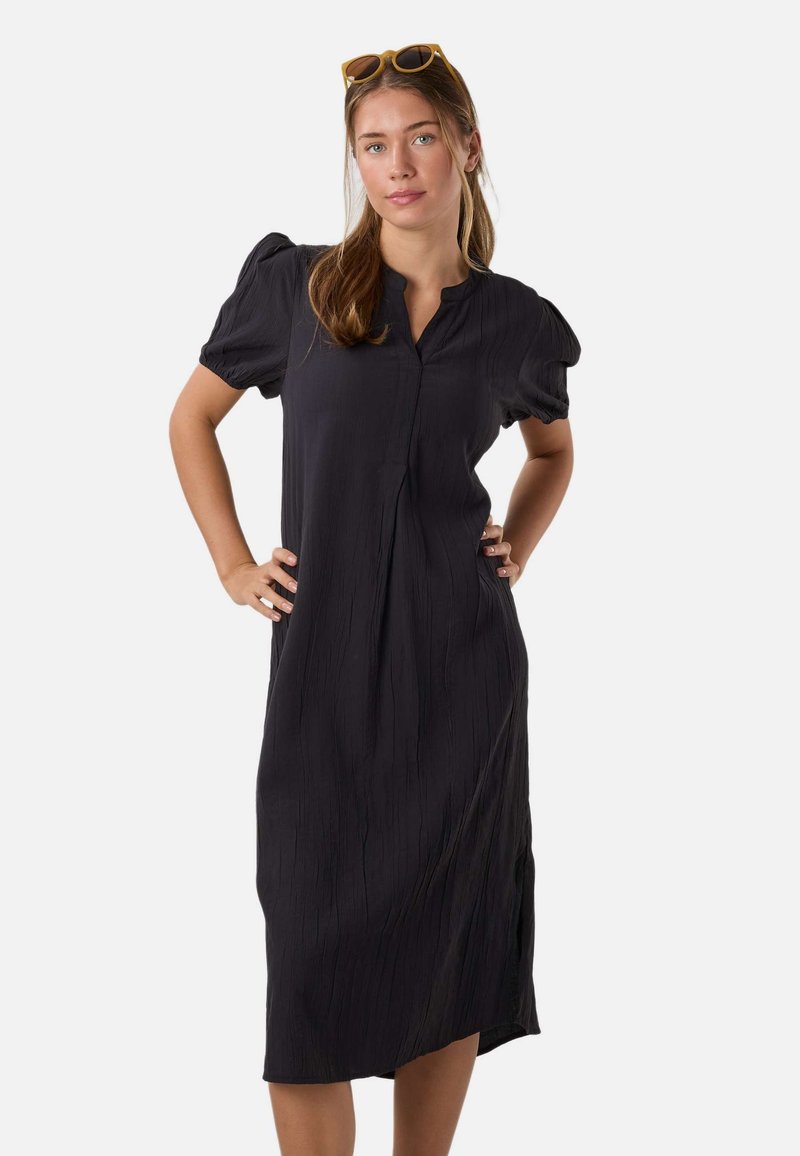 Femme aux longs cheveux portant une robe midi noire à manches bouffantes, les mains sur les hanches, avec des lunettes de soleil jaunes sur la tête, sur un fond uni.