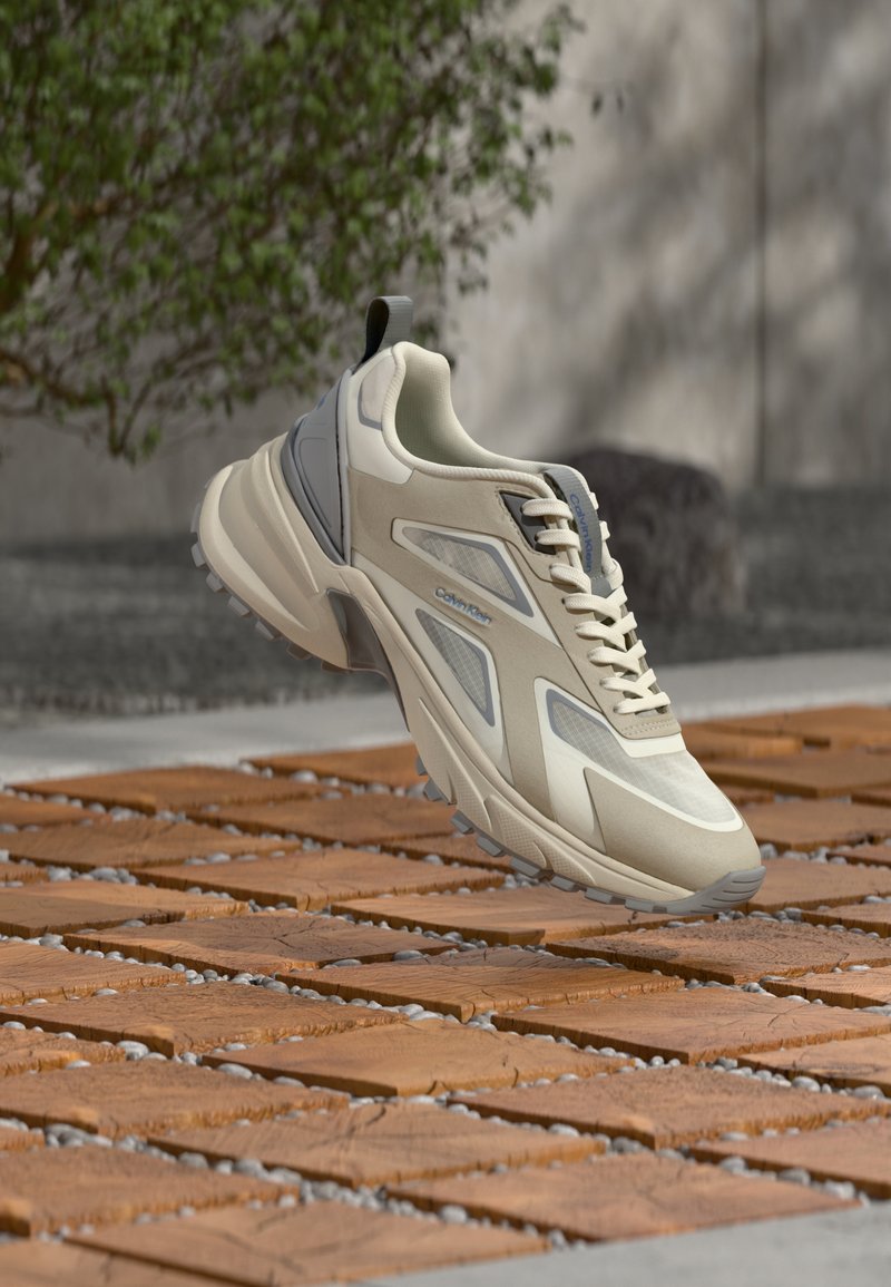 Μπεζ και γκρι αθλητικό παπούτσι με υφασμένη σόλα, κορδόνια και λογότυπο Calvin Klein, τοποθετημένο πάνω σε ξύλινη και χαλικώδη πλακόστρωση σε εξωτερικό χώρο.