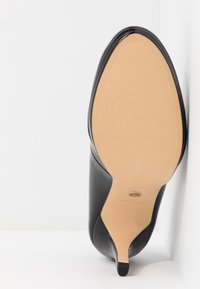 Chaussure femme à talon haut en cuir noir vue par la semelle, fond beige, bout pointu et talon fin sur fond blanc.