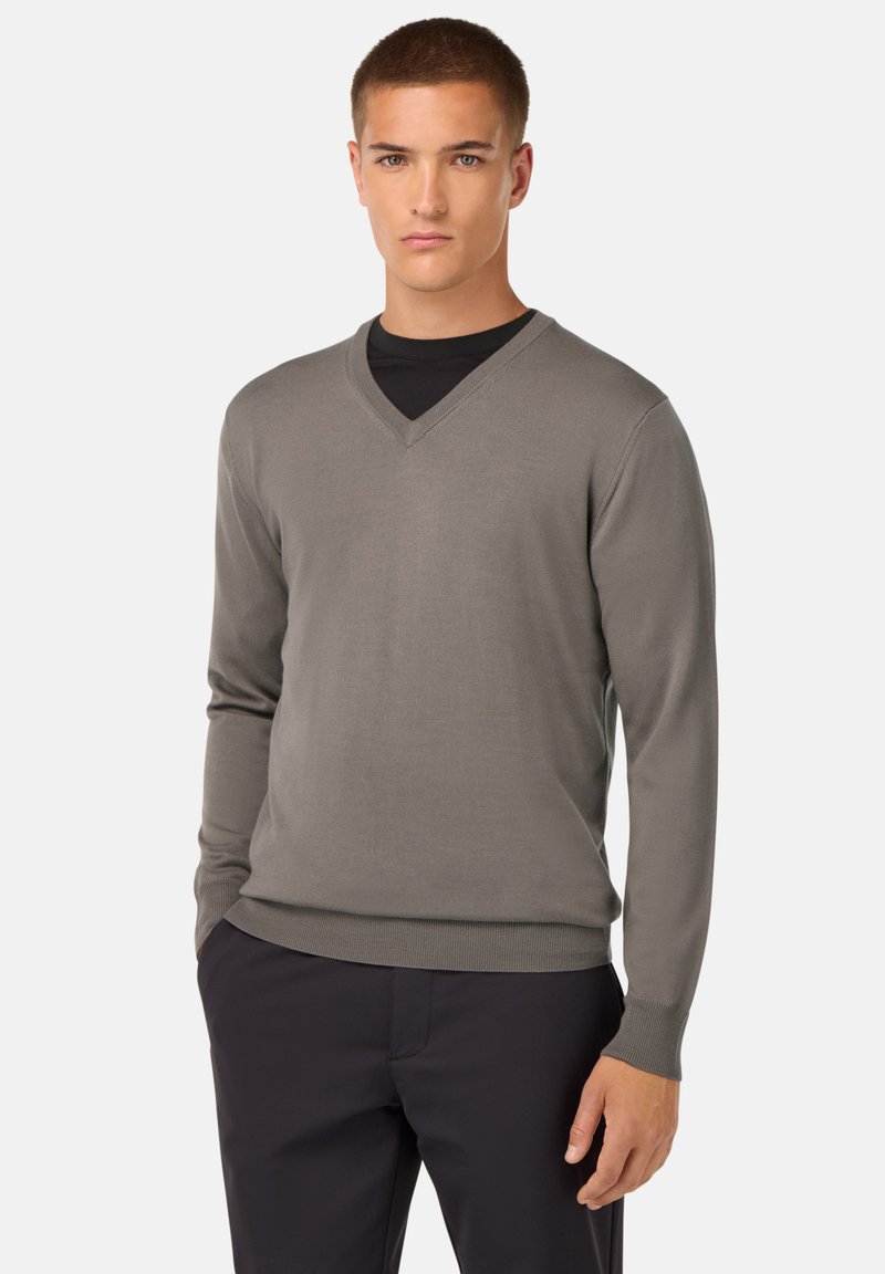 V-Ausschnitt-Pullover in Olivgrün, aus weichem Strickmaterial, schlanker Schnitt, lange Ärmel, gerippte Bündchen und Saum, getragen über einem schwarzen Hemd.