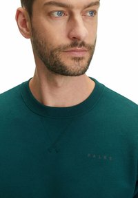 Felpa verde con collo rotondo, polsini a costine e un logo sottile sul petto. Il tessuto appare morbido con una consistenza liscia.