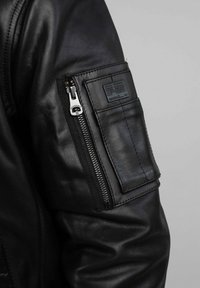 Veste en cuir noir avec une poche à fermeture éclair sur la manche, texture lisse et des détails de couture bien visibles. Quincaillerie de fermeture éclair visible sur la poche.