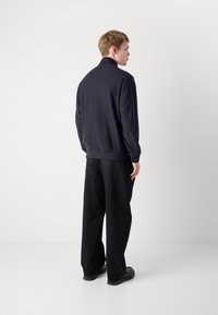 Jonge man die zijwaarts staat, gekleed in een donkerblauwe hoge hals trui, zwarte wijde broek en zwarte schoenen, tegen een witte achtergrond.
