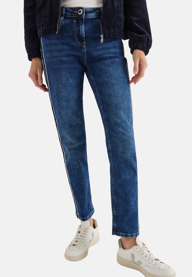 Vaqueros de mezclilla azules con un corte recto, con rayas laterales y cierre de cremallera. Combinados con zapatillas blancas para un look casual.
