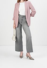 Blazer rose clair, t-shirt blanc, jean gris taille haute à jambes larges, et escarpins beiges à bouts pointus, tenant un petit sac à main blanc.