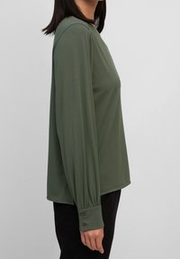 Maglietta a maniche lunghe, verde oliva, con scollatura tonda e polsini arricciati e con bottoni; realizzata in tessuto morbido e liscio, presenta una vestibilità comoda.