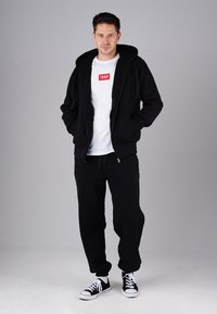 Man wearing black hoodie, black sweatpants, black and white sneakers, and white t-shirt with red logo, standing against gray background.