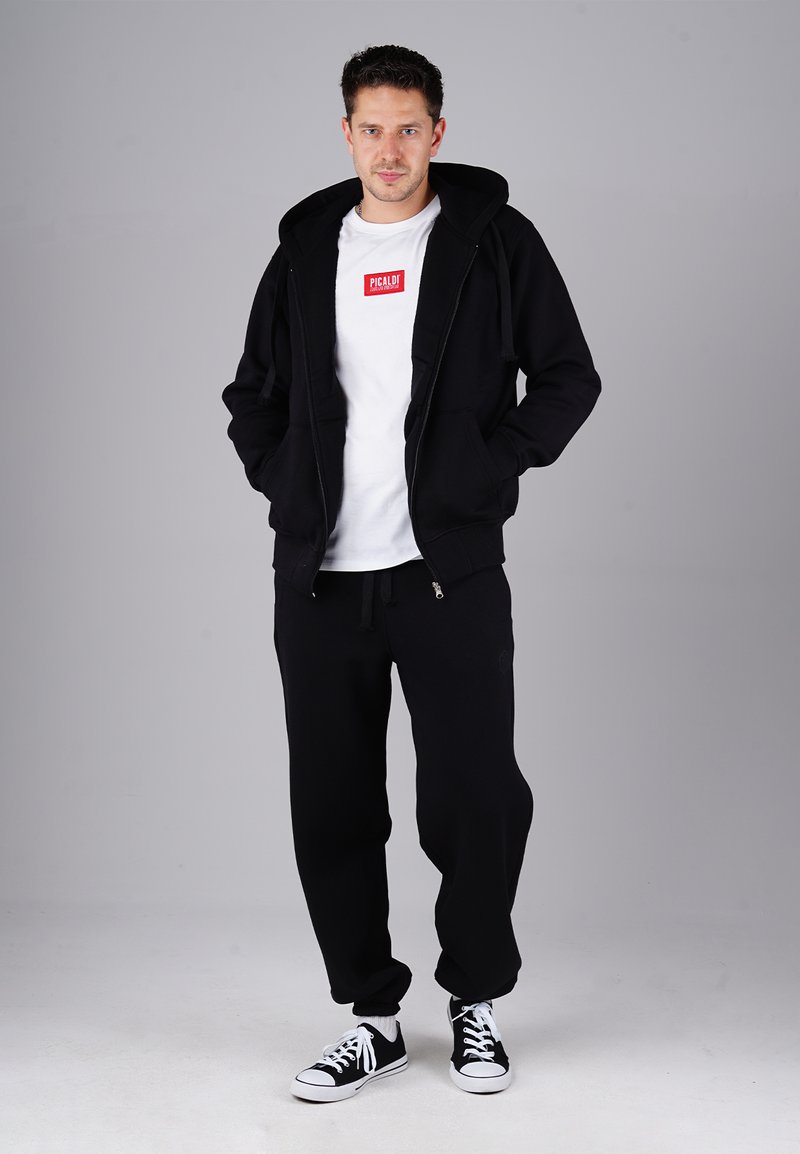 Man wearing black hoodie, black sweatpants, black and white sneakers, and white t-shirt with red logo, standing against gray background.
