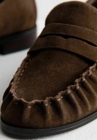 Scarpa moccasin in suede marrone con dettagli cuciti e suola nera su una superficie chiara.