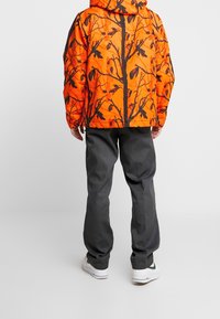 Giacca arancione imbottita con motivo di rami neri, abbinata a pantaloni grigio scuro e sneakers bianche, vista da dietro.