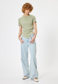 Lys grøn tætsiddende T-shirt med rund hals og korte ærmer, parret med højtaljede lys blå flagrende jeans og beige flade sko.