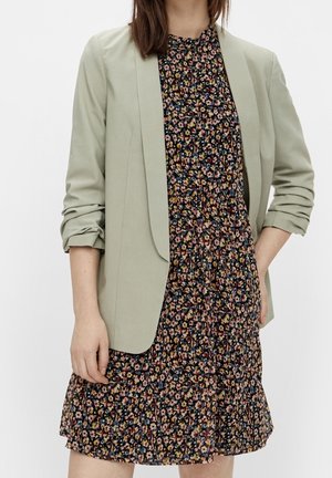 Kvinde iført en lys salviegrøn blazer med rynkede ærmer over en sort blomsterkjole med små flerfarvede blomster.