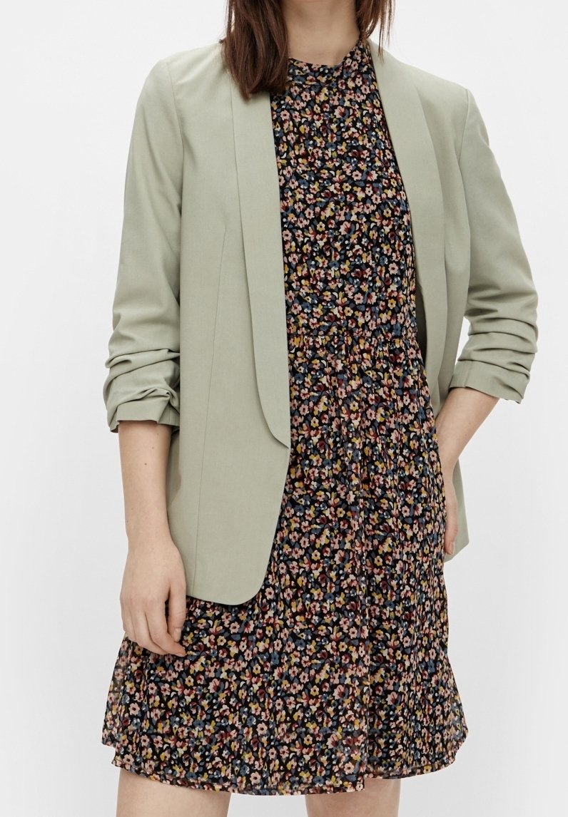 Femme portant un blazer vert sauge clair avec des manches froncées sur une robe noire à petites fleurs multicolores.