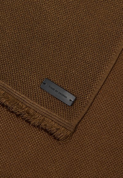 Cobertor castanho texturizado com bordas desfiadas, apresentando uma etiqueta preta em relevo com "TIGER OF SWEDEN." Feito de material tecido.