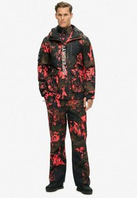 Traje de invierno impermeable en negro con patrones de camuflaje en rojo y marrón oscuro. Cuenta con capucha, múltiples bolsillos y cremalleras de alta resistencia.