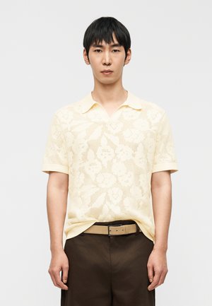 Νέος άνδρας με ανοιχτό κίτρινο πλεκτό πουκάμισο με λουλουδένιο μοτίβο και κοντά μανίκια, μπουκαφτωμένο σε σκούρο καφέ παντελόνι με μπεζ ζώνη.