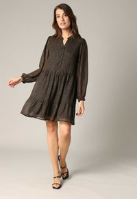 Bruine jurk met een patroon, kraag, lange mouwen en een uitlopende rok. Transparante, gestructureerde stof met plooien, gedragen met zwarte sandaaltjes met bandjes.