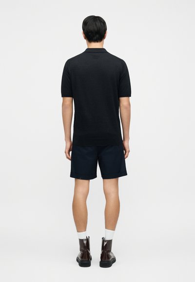 Polo noir à manches courtes en tissu lisse, associé à un short bleu marine, des chaussettes blanches et des bottines marron foncé.