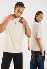 Beige Baumwoll-T-Shirts mit kurzen Ärmeln, ausgestattet mit einem kleinen schwarzen Schlangendesign. Tragen von zwei Modellen, wobei eines nach vorne und das andere nach hinten blickt.