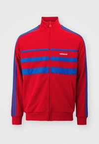 Červená sportovní bunda na zip s modrými pruhy přes hrudník a rukávy, s vyšívaným logem adidas na levé straně.