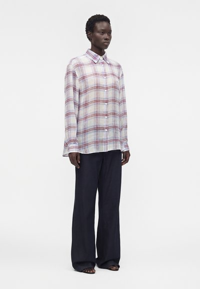 Άτομο με σκούρο δέρμα που φοράει άνετο λευκό πουκάμισο με κόκκινο και μπλε καρό και φαρδιές μαύρες παντελόνες, στέκεται μπροστά σε λευκό φόντο.