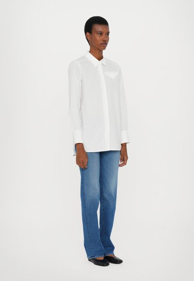 Camicia bianca con colletto a punta e taschino sul petto, abbinata a jeans blu a gamba larga e ballerine nere. Tessuto morbido, vestibilità casual.