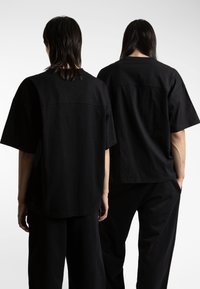 Black, oversized cotton t-shirts with short sleeves, featuring a simple design and a horizontal seam across the back. Worn with black pants.