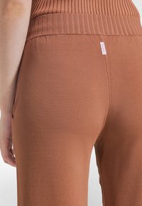 Primer plano de una persona que lleva pantalones de punto acanalado marrón de talle alto con una pequeña etiqueta rosa de DEHA en la parte posterior de la cintura.