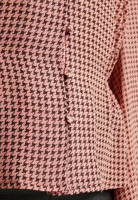 Růžový a hnědý houndstooth vzorovaný materiál s mírně texturovaným povrchem. Obsahuje knoflíky podél švu pro zapínání.