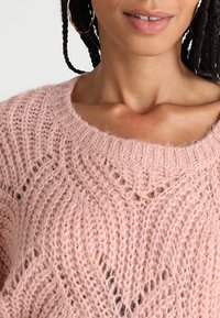Jasnoróżowy sweter robiony na drutach o luźnym kroju, szerokim dekolcie i delikatnym, wzorzystym wykończeniu z małymi wycięciami.