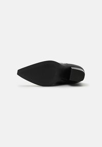 Tacón de cuero negro con punta afilada y suela de goma texturizada. Presenta un acabado brillante y un tacón de altura estándar para mayor estabilidad.