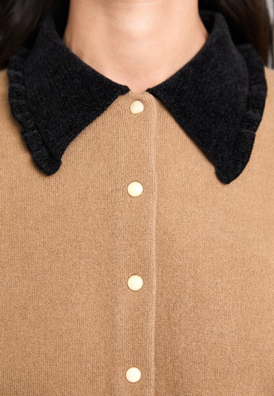 Rudas marškinių tipo drabužis su tekstūruotu juodu apkakle, volangais ir kreminiais mygtukais, pagamintas iš minkštos megztos medžiagos.