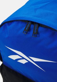 Mochila azul com fundo preto, tecido liso, fecho de duas zíperes com puxadores da Reebok e um grande logótipo branco.