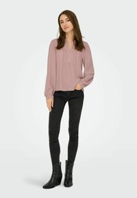 Blusa rosa chiaro a maniche lunghe con scollo a V e dettaglio plissè sul davanti, abbinata a jeans skinny neri e stivaletti neri. Texture liscia.