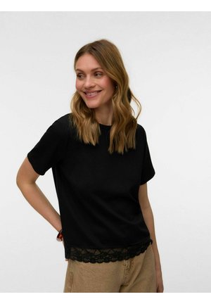 Kvinde med bølget blondt hår smiler, iført en sort kortærmet top med blondekant og beige bukser mod en ensfarvet hvid baggrund.