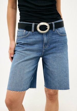 Persoon draagt halflange blauwe denimshorts met rafelige zoom en een zwarte riem met een grote ovale zilveren gesp.