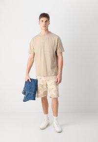 T-shirt beige à col rond avec graphique subtil, shorts camouflés beiges, et baskets blanches. Tenant une veste en denim bleu. Fond neutre.