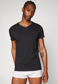 Black cotton v-neck t-shirt with short sleeves, worn by a model. White boxer briefs visible. Simple design, smooth texture, fitted shape.