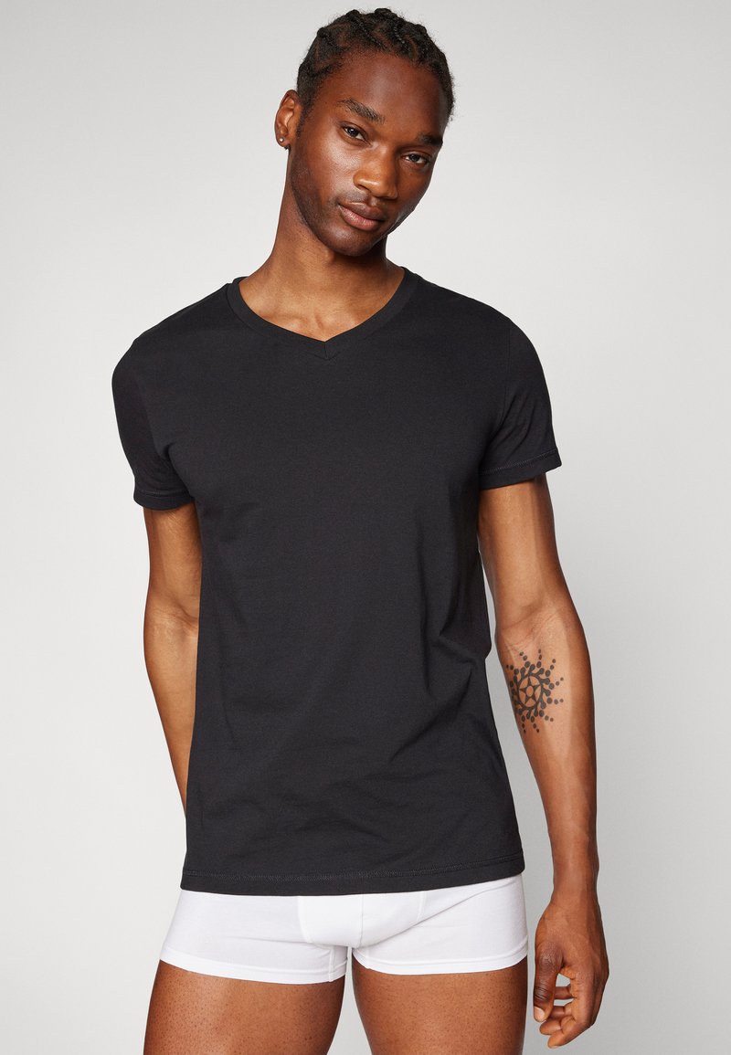 Schwarzes Baumwoll-T-Shirt mit V-Ausschnitt und kurzen Ärmeln, getragen von einem Model. Weiße Boxershorts sichtbar. Einfaches Design, glatte Textur, figurbetonte Form.