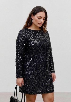 Mujer con cabello largo y oscuro, vestida con un vestido negro de lentejuelas de manga larga, sosteniendo un bolso negro, mirando hacia abajo sobre un fondo claro y liso.