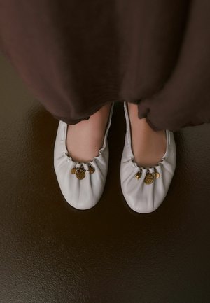 Vita läderballerinas med rynkade kanter, prydda med guldiga berlocker på tån. Slät textur och rundad form.